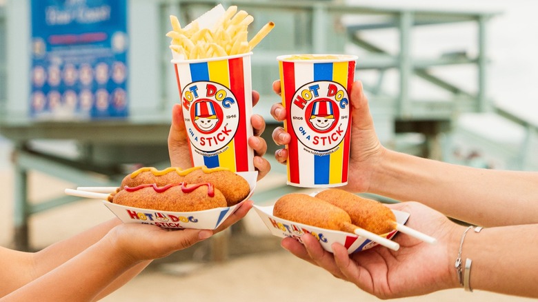 Hands holding corndogs, a drink, and a cup of fries from hot dog on a stick