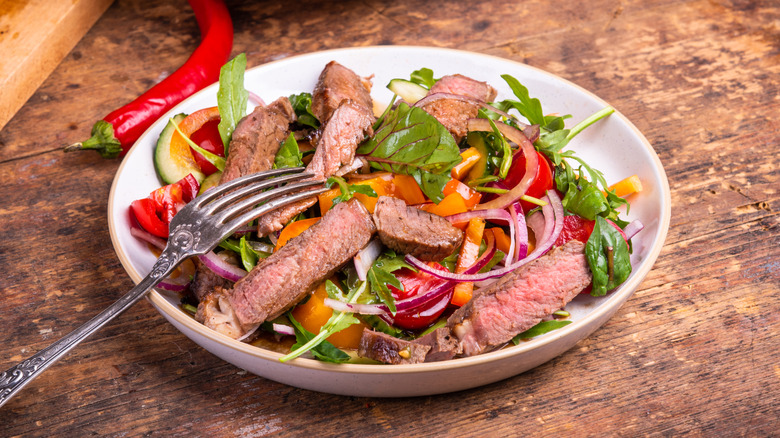 Plate of salad topped with steak pieces on a wooden table with a fork and a chili pepper on the side