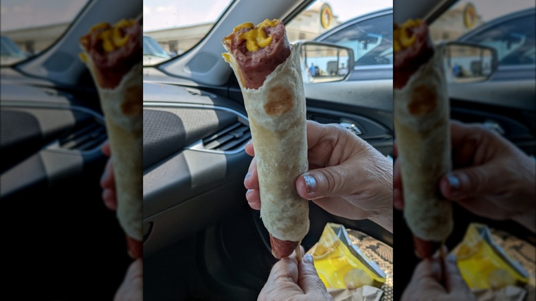 Hand holding a sausage on a stick wrapped in a tortilla with a bite taken out of it