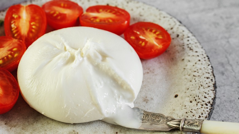 Whole burrata surrounded by tomatoes on a black and white plate–sliver knife next to it