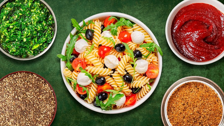 Pasta salad dish surrounded by various toppings against a green background
