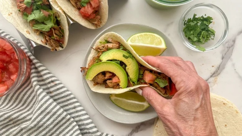 View from above of a hand holding jackfruit carnitas packed with tomatoes, avocado, and herbs with limes on the side