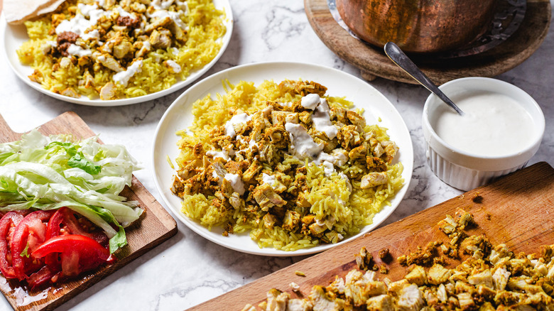 Two plates of chicken and rice with lettuce and tomato on the side, rice and more chicken chopped on board