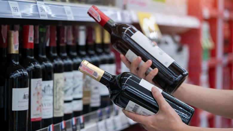A person holds two bottles of wine in front of a shelf of wine