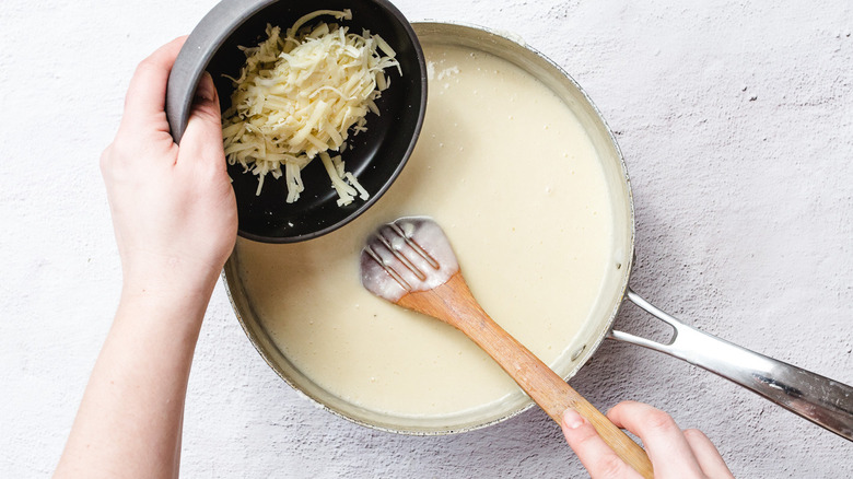 Hand pouring bowl of shredded cheese into saucepan with creamy sauce