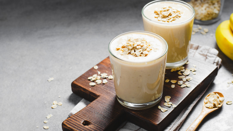 Two pale oat smoothies on a board surrounded by oats with bananas in the background