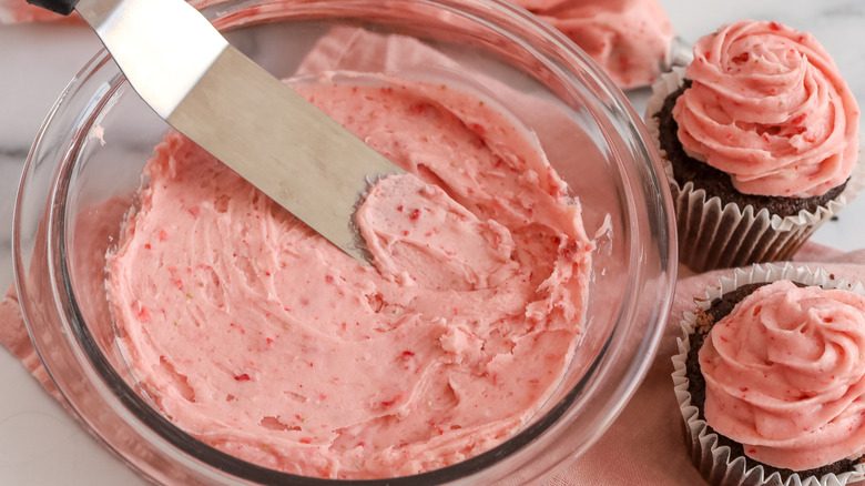 pink frosting in a clear bowl next to two pink-frosted chocolate cupcakes