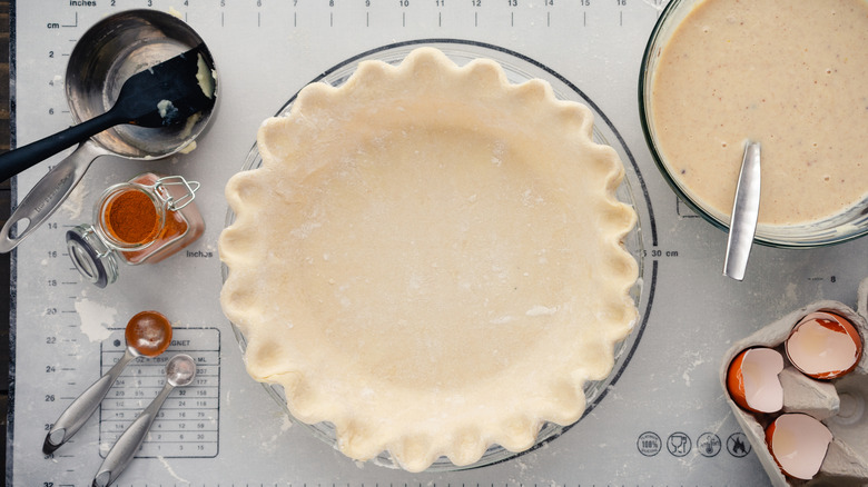 Raw pie crust surrounded by other baking supplies.