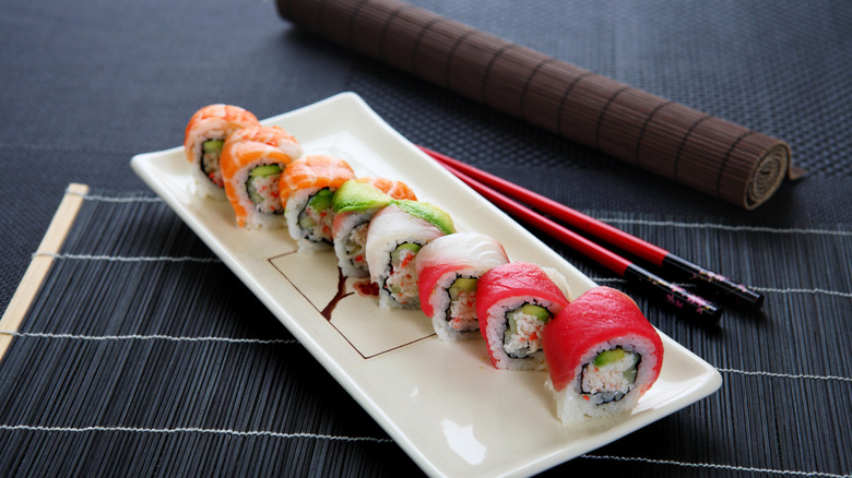 A rainbow roll on a plate sitting on a bamboo mat next to a pair of chopsticks