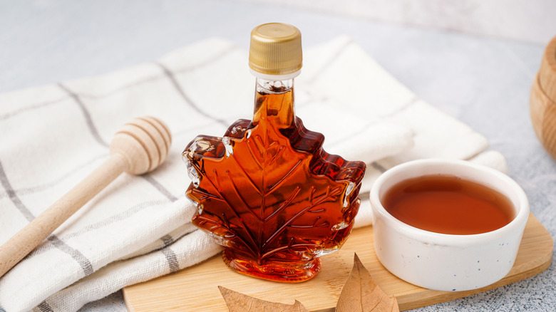 A maple leaf shaped glass bottle of syrup next to a ramekin of syrup, a maple leaf, a towel, and a honey dipper